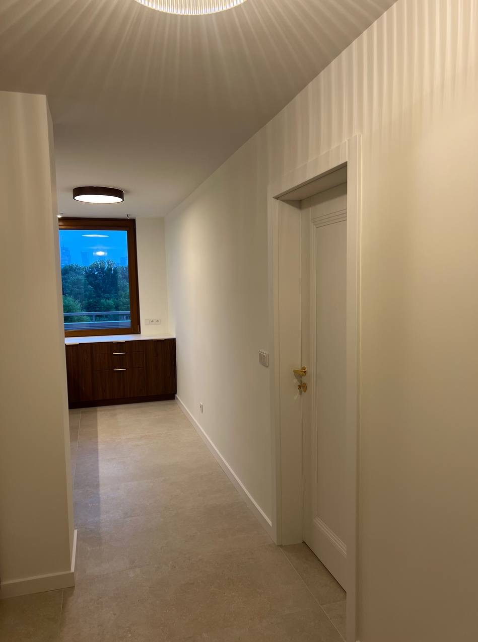 Jasne wnętrze po remoncie: kremowe ściany, beżowa podłoga, białe drzwi z złotymi klamkami i zabudowa okienna z ciemnego drewna. Nowoczesne oświetlenie sufitowe.