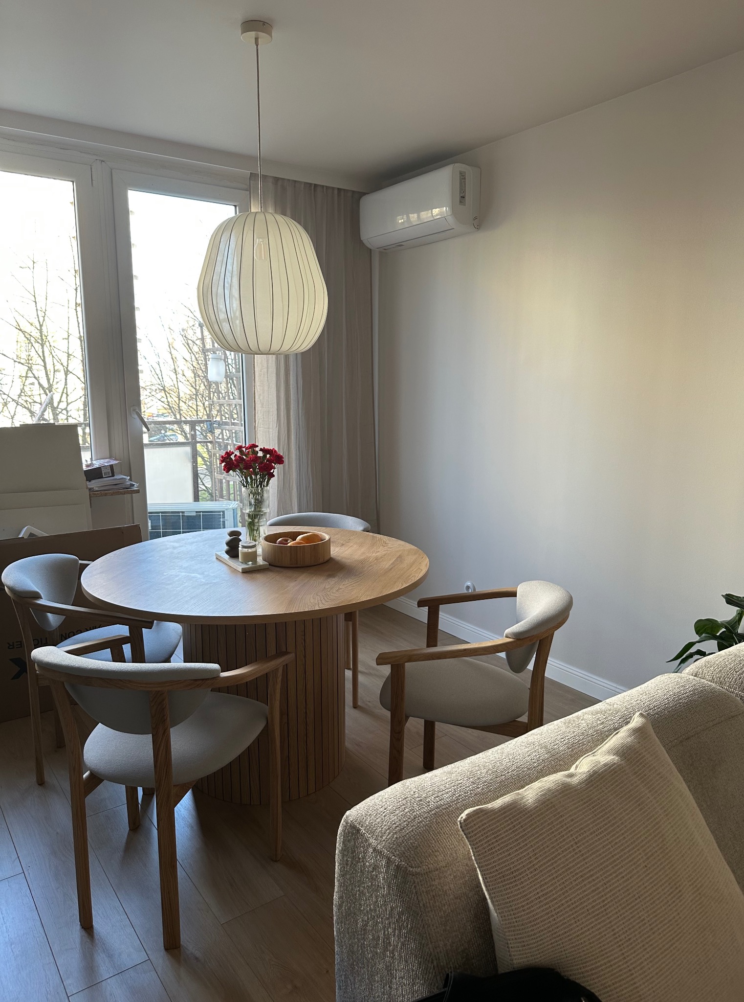 Jasne wnętrze jadalni z okrągłym stołem z drewna i czterema krzesłami. Na stole wazon z czerwonymi kwiatami i dekoracje. Widoczny fragment sofy i klimatyzator.