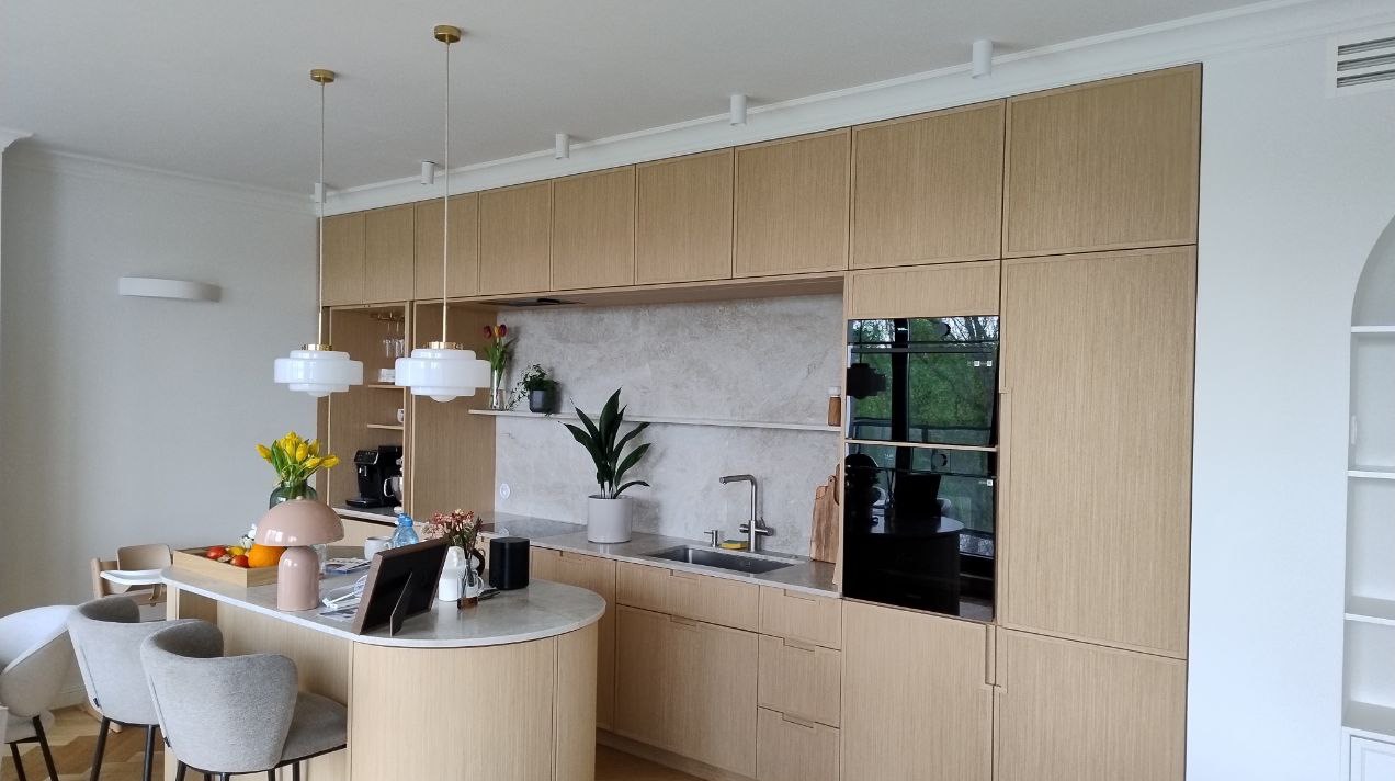 Jasna, nowoczesna kuchnia z drewnianymi szafkami, wyspą i oświetleniem punktowym. Widoczne sprzęty AGD i dekoracje roślinne. Minimalistyczny design.