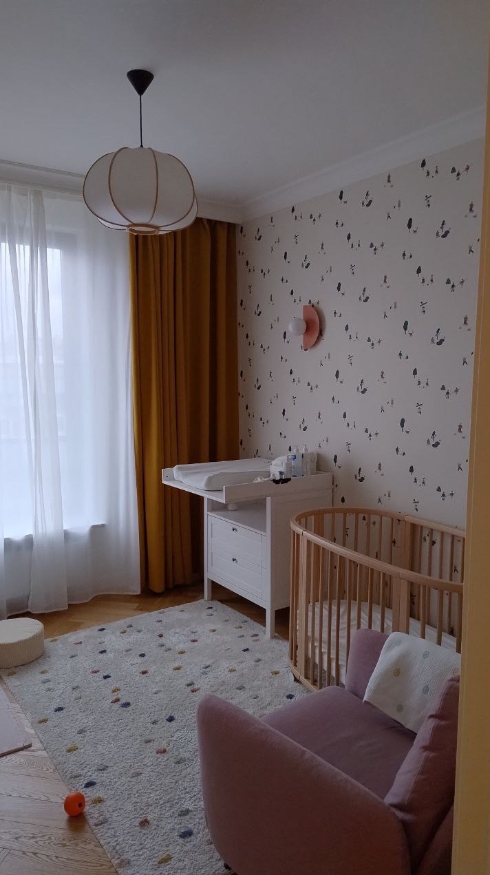 Jasny pokój dziecięcy z okrągłym łóżeczkiem, komodą z przewijakiem, wzorzystą tapetą i dywanem w kropki. Złote zasłony i lampa dodają ciepła wnętrzu.