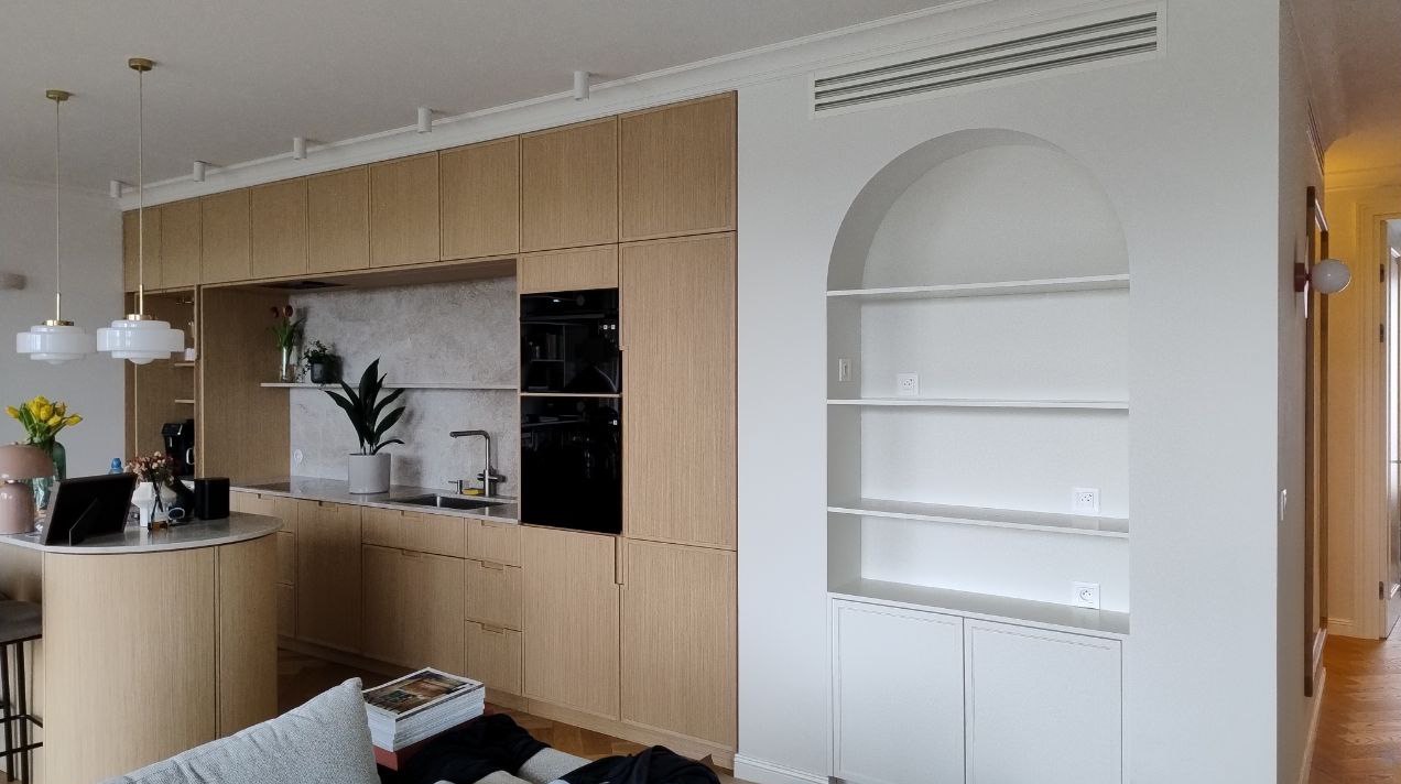 Nowoczesna kuchnia z drewnianymi szafkami i wyspą, uzupełniona o wnękę z półkami w kształcie łuku. Minimalistyczny design, jasne kolory, eleganckie oświetlenie.