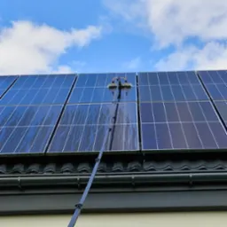 Czyszczenie paneli słonecznych na dachu domu za pomocą szczotki na długim wysięgniku. Widoczne smugi wody na panelach, błękitne niebo z chmurami w tle.