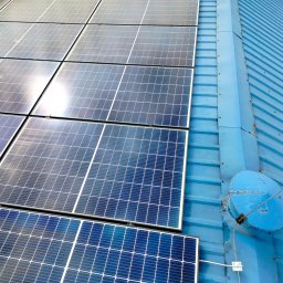 Adam Frankiewicz BŁYSK - ZYSK - Błękitny dach z panelami fotowoltaicznymi i anteną satelitarną. Widok z góry, ukazujący regularny układ paneli i detale konstrukcji dachu.