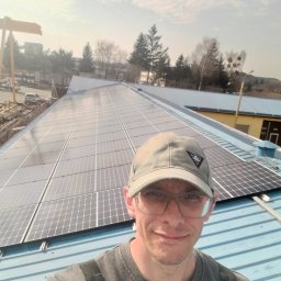 Adam Frankiewicz BŁYSK - ZYSK - Instalator w okularach na dachu z panelami słonecznymi, widok z góry na instalację fotowoltaiczną w słoneczny dzień, w tle drzewa i budynki przemysłowe.