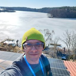 Adam Frankiewicz BŁYSK - ZYSK - Selfie instalatora na dachu z panelami słonecznymi, w tle zamarznięte jezioro i las. Widoczny pracownik w zielonej czapce i okularach, z identyfikatorem firmy na szyi.