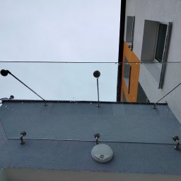 Adam Frankiewicz BŁYSK - ZYSK - Szklany daszek nad wejściem do budynku, mocowany na metalowych wspornikach. Widoczne krople deszczu na szkle i elewacja budynku w tle.