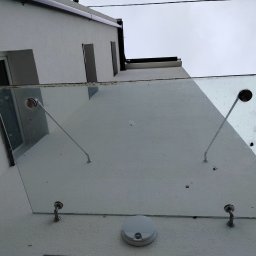 Adam Frankiewicz BŁYSK - ZYSK - Szklany daszek nad wejściem do budynku, widok z dołu. Widoczne mocowania i zabrudzenia na szkle. Biała elewacja budynku i fragment nieba w tle.