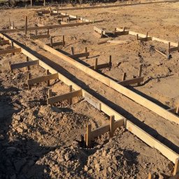 ASAMI Paweł Kogut - Szczegółowy widok szalunków fundamentowych z desek na piaszczystym gruncie, przygotowanych pod wylanie betonu. Widoczne drewniane podpory i elementy konstrukcyjne.