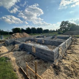 ASAMI Paweł Kogut - Wzniesione ściany fundamentowe z szarego bloczka betonowego, z widocznymi prętami zbrojeniowymi i drewnianymi szalunkami, na tle błękitnego nieba z chmurami.