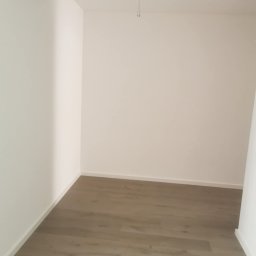 Bartłomiej Powietrzyński - Pusty pokój po remoncie: świeżo pomalowane białe ściany, ułożona podłoga z paneli w odcieniach szarości. Widoczny fragment kabla wystający z sufitu.