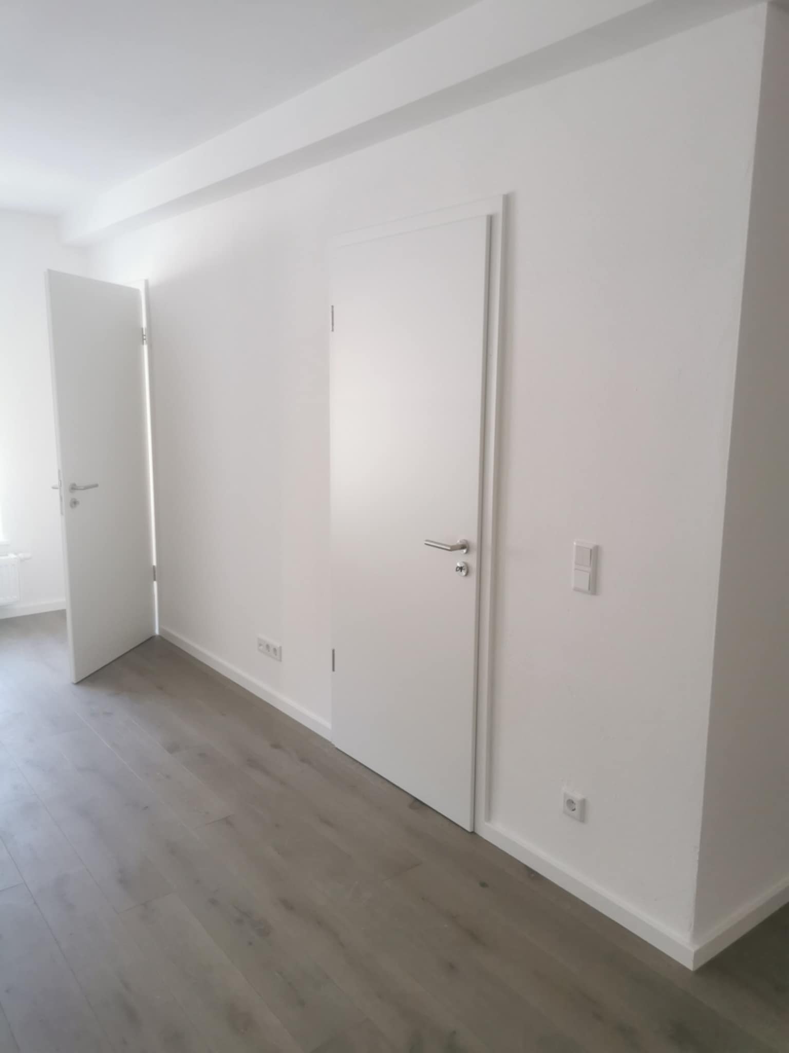 Jasne wnętrze po remoncie: białe ściany, szara podłoga z paneli, dwoje białych drzwi z srebrnymi klamkami, gniazdka elektryczne. Minimalistyczny design.