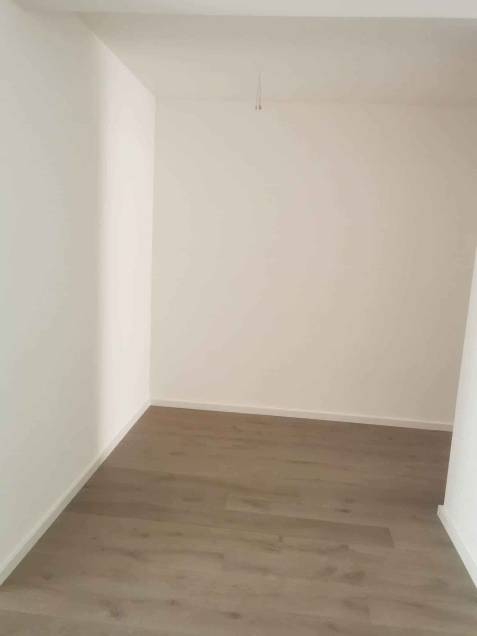 Pusty pokój po remoncie: świeżo pomalowane białe ściany, ułożona podłoga z paneli w odcieniach szarości. Widoczny fragment kabla wystający z sufitu.