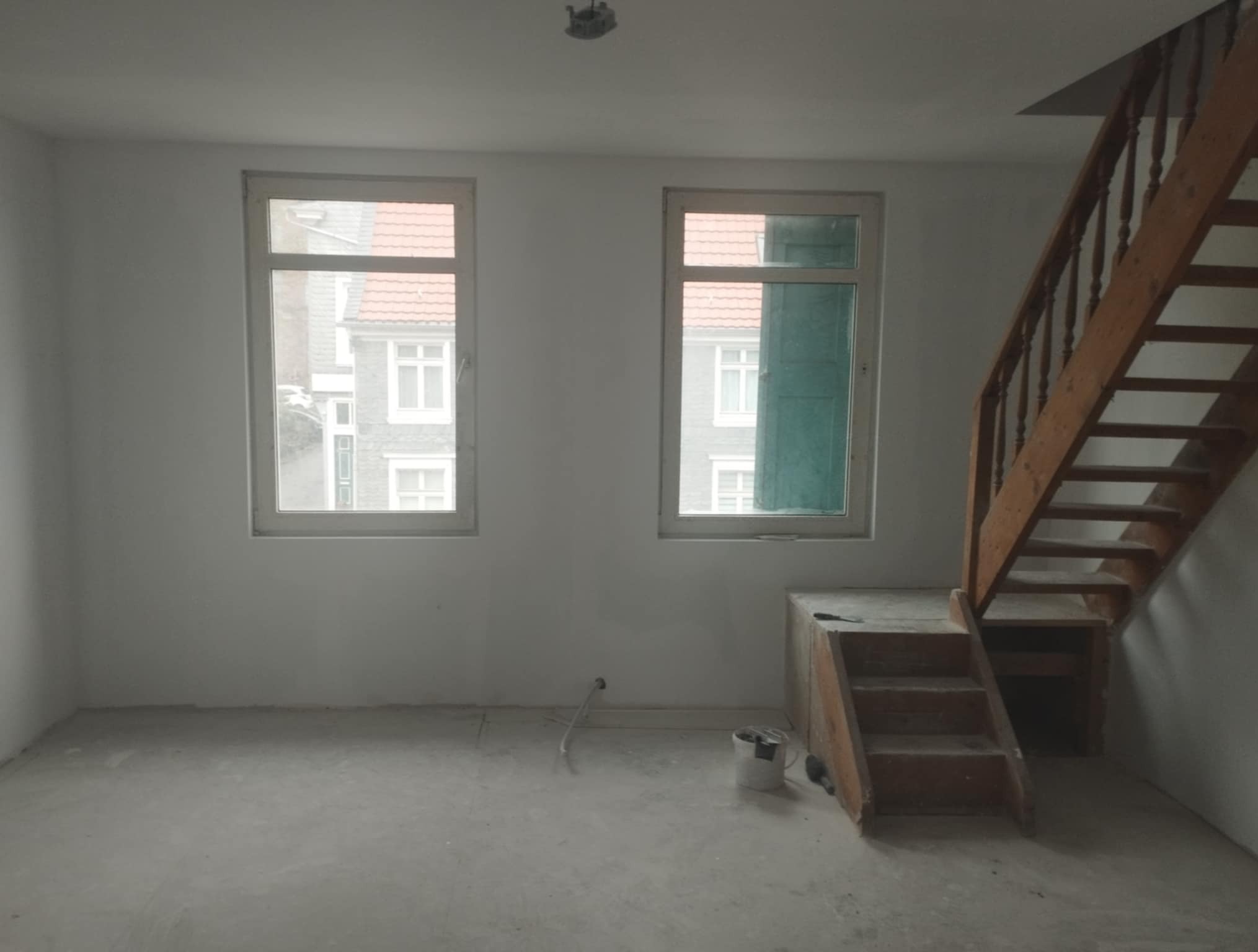 Wnętrze pokoju w trakcie prac wykończeniowych: białe ściany, drewniane schody, dwa okna z widokiem na budynek. Na podłodze widoczne ślady prac, stoi wiadro z farbą.