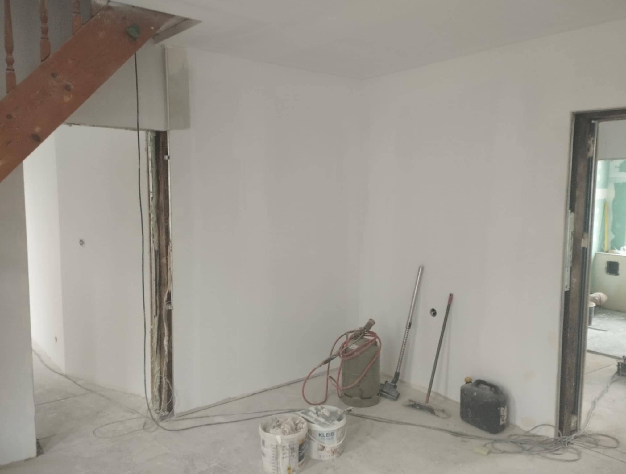 Wnętrze w trakcie szpachlowania: gładkie, białe ściany, widoczne narzędzia (szpachelka, agregat), wiadro z masą szpachlową. Fragment schodów i ościeżnica drzwi.