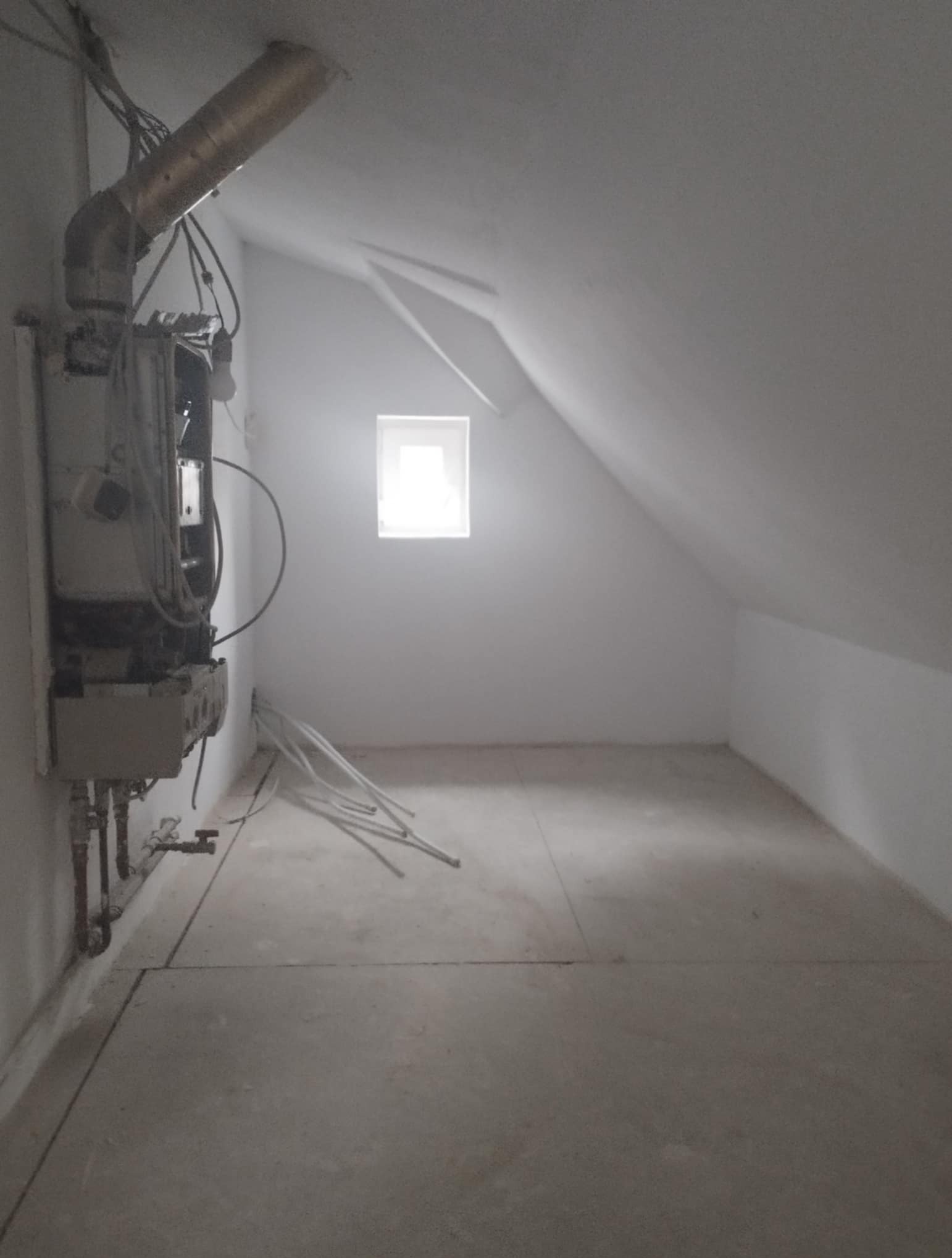 Poddasze ze skośnym sufitem w trakcie prac wykończeniowych. Widoczny piec gazowy, rury i kable. Małe okno wpuszcza jasne światło.