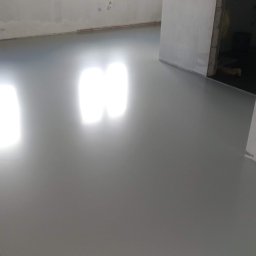 InterioDecor - dekoracyjne posadzki żywiczne - Gładka, szara posadzka w surowym wnętrzu, odbijająca światło okien. Widoczne niedokończone ściany i przejście do innego pomieszczenia. Minimalistyczny, nowoczesny wygląd.