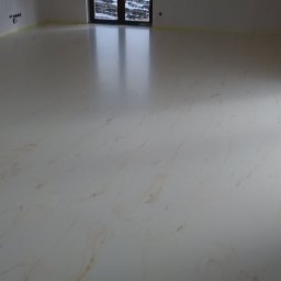 InterioDecor - dekoracyjne posadzki żywiczne - Jasna, błyszcząca posadzka z delikatnym wzorem marmuru w przestronnym wnętrzu. Widoczne okna i fragmenty ścian z taśmą malarską zabezpieczającą.