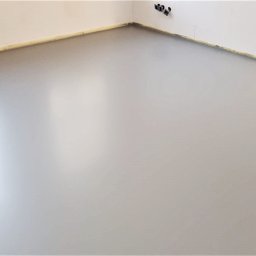 InterioDecor - dekoracyjne posadzki żywiczne - Jasnoszara, gładka posadzka w rogu pomieszczenia z grzejnikiem i gniazdkami. Widoczne taśmy zabezpieczające przy ścianach. Minimalistyczny design, równa powierzchnia.