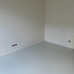 InterioDecor - dekoracyjne posadzki żywiczne - Jasnoszara, gładka posadzka w rogu pomieszczenia z białymi ścianami. Widoczne trzy otwory na kable w ścianie i jeden otwór z kablem. Minimalistyczne wykończenie wnętrza.