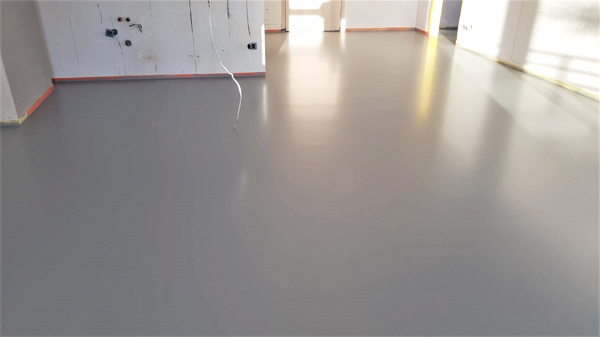 Szara, gładka posadzka w jasnym wnętrzu z przygotowanymi ścianami do malowania. Widoczne kable i taśmy zabezpieczające. Minimalistyczny design.