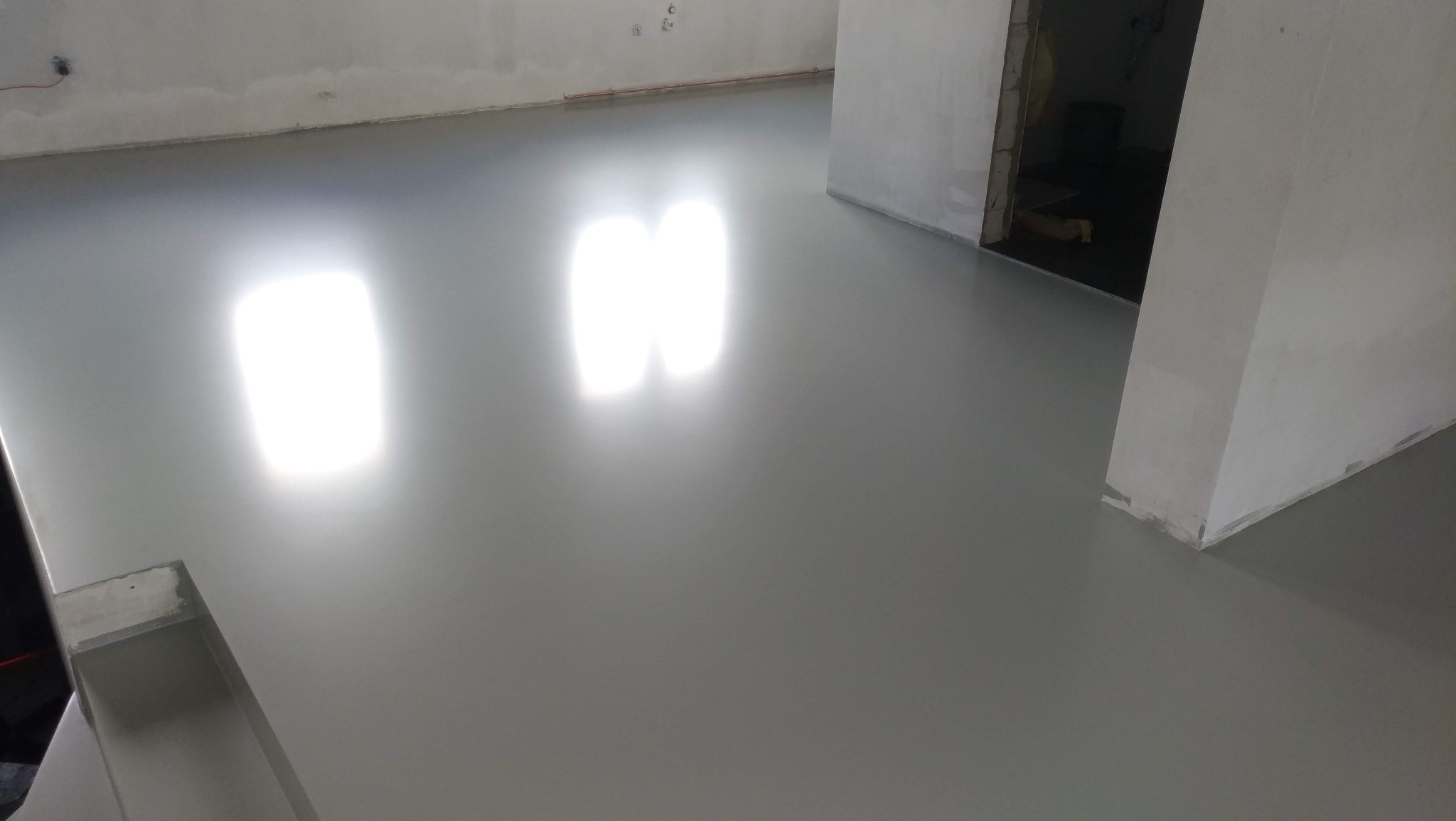 Gładka, szara posadzka w surowym wnętrzu, odbijająca światło okien. Widoczne niedokończone ściany i przejście do innego pomieszczenia. Minimalistyczny, nowoczesny wygląd.