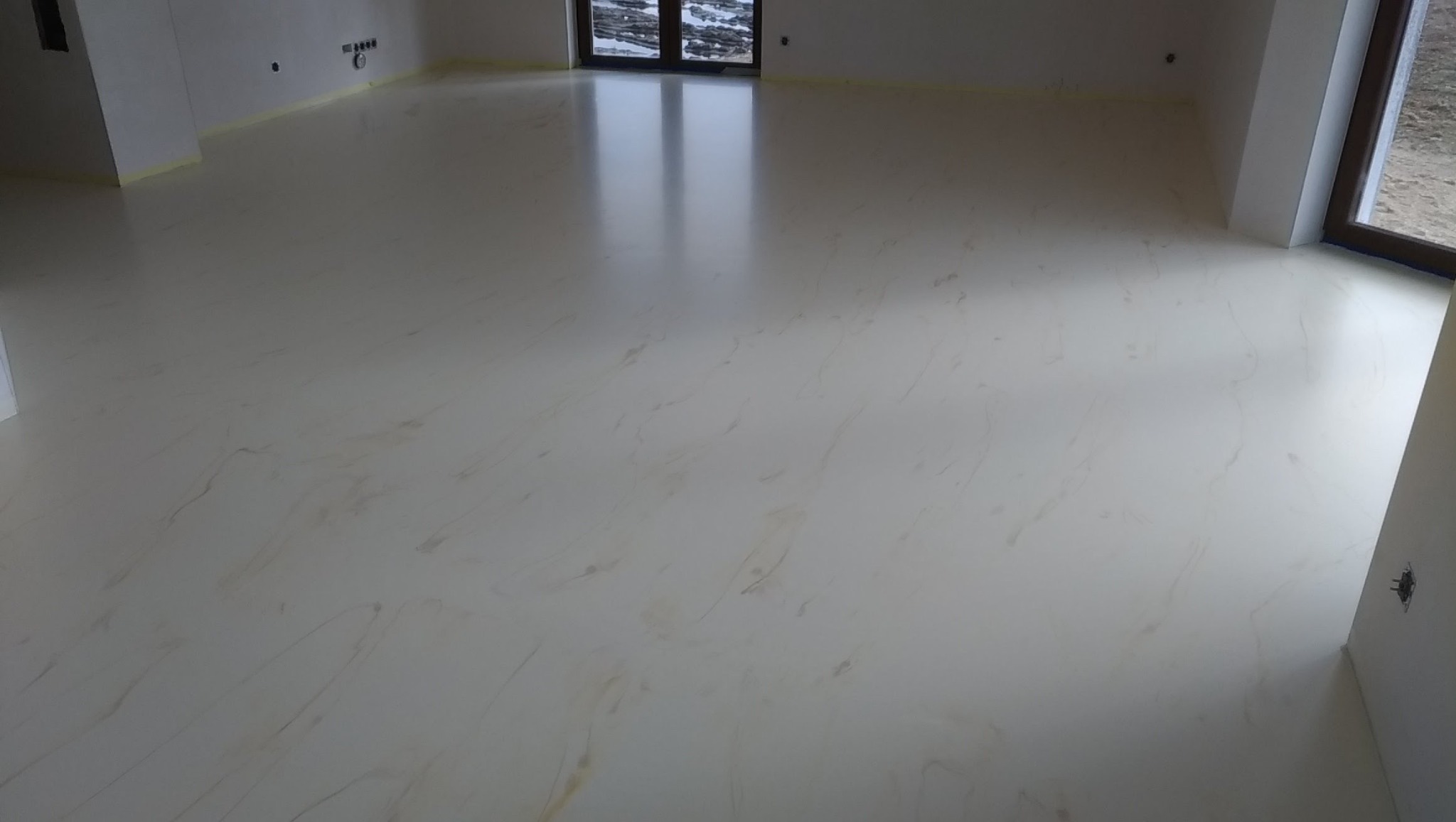 Jasna, błyszcząca posadzka z delikatnym wzorem marmuru w przestronnym wnętrzu. Widoczne okna i fragmenty ścian z taśmą malarską zabezpieczającą.