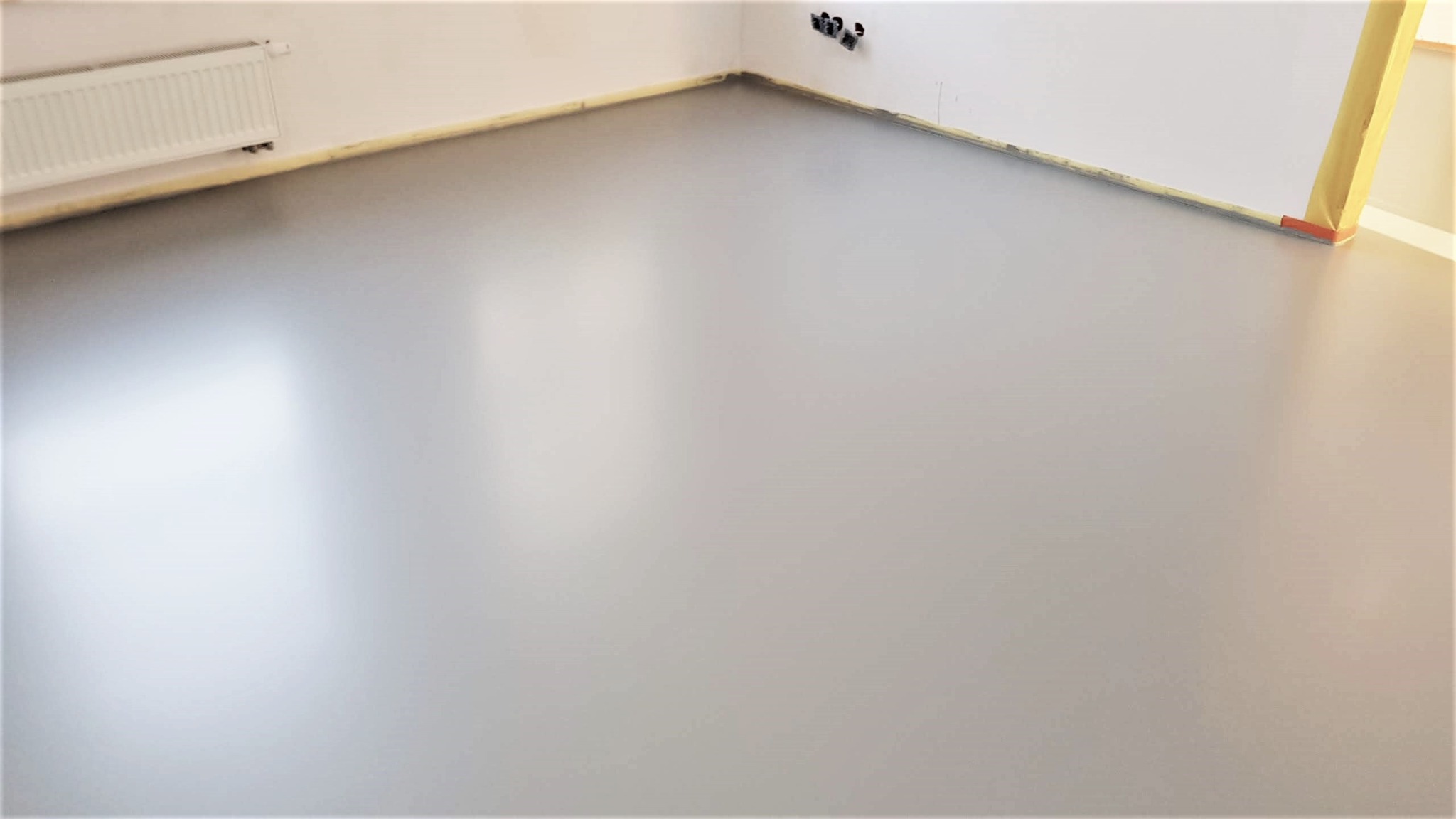 Jasnoszara, gładka posadzka w rogu pomieszczenia z grzejnikiem i gniazdkami. Widoczne taśmy zabezpieczające przy ścianach. Minimalistyczny design, równa powierzchnia.