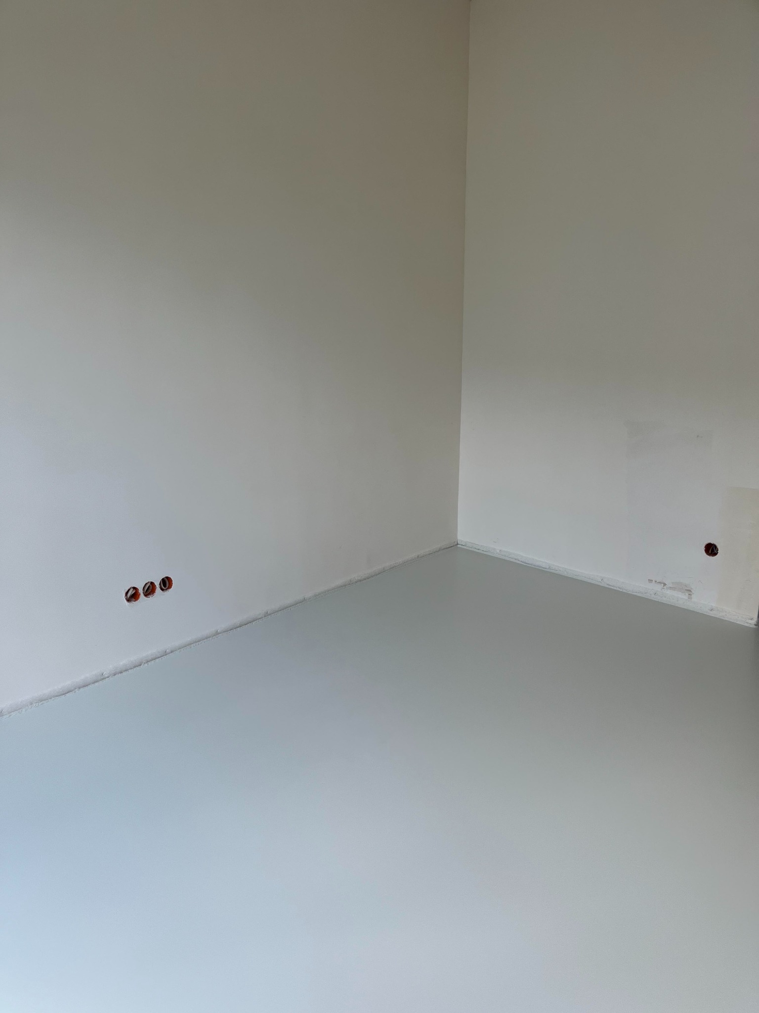 Jasnoszara, gładka posadzka w rogu pomieszczenia z białymi ścianami. Widoczne trzy otwory na kable w ścianie i jeden otwór z kablem. Minimalistyczne wykończenie wnętrza.