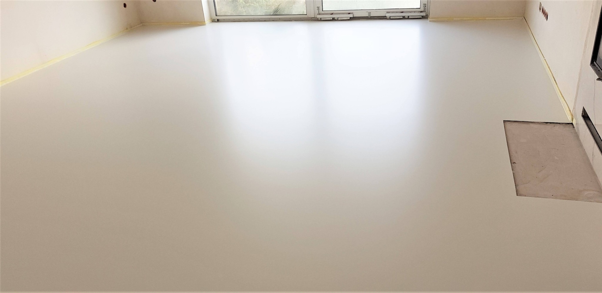 Jasna, gładka posadzka w pokoju z oknem, zabezpieczona taśmą malarską przy ścianach. Widoczny fragment ściany z otworem instalacyjnym.