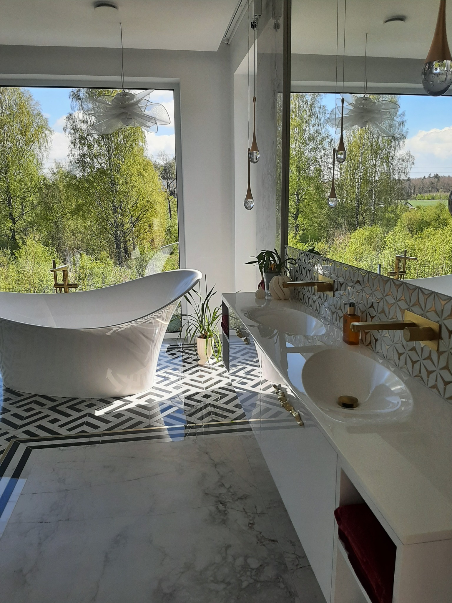 Nowoczesna łazienka z wolnostojącą wanną na tle okna z widokiem na zieleń. Geometryczny wzór płytek podłogowych i złote akcenty armatury dodają elegancji.