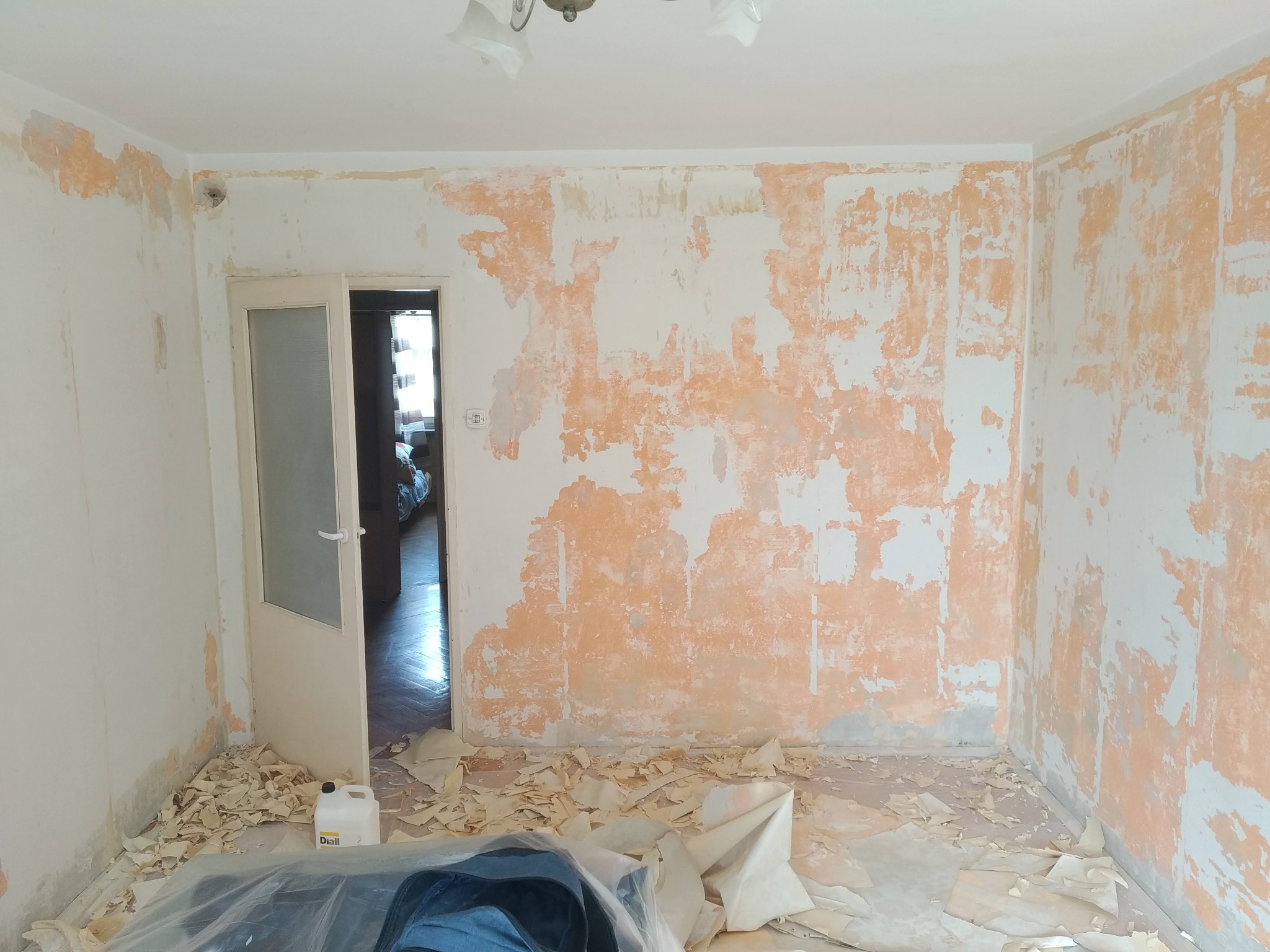 Pokój w trakcie przygotowania do malowania: ściany ze śladami starej farby, resztki tapet na podłodze, otwarte drzwi do kolejnego pomieszczenia.