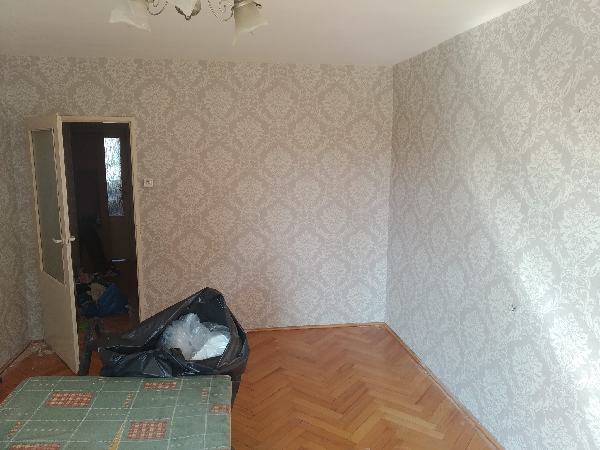 Pokój po malowaniu ścian z wzorzystą tapetą w odcieniach szarości. Na podłodze parkiet w jodełkę i czarny worek na śmieci. Widoczne drzwi do innego pomieszczenia.