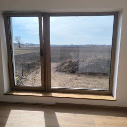 Yana P. - Duże okno z drewnianą ramą, widok na pole i niebo. Na parapecie leżą drobne elementy. Na podłodze widoczne promienie słoneczne.