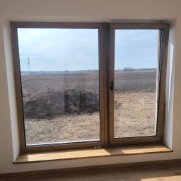 Yana P. - Drewniane okno z widokiem na pole w słoneczny dzień. Na parapecie leży mała szczotka. Podłoga z paneli odbija promienie słoneczne.