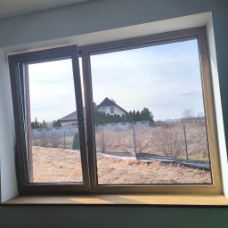 Yana P. - Drewniane okno z widokiem na ogród i dom. Jedna część okna jest uchylona, widać krajobraz za oknem w słoneczny dzień. Na parapecie widoczne refleksy światła.