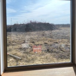 Yana P. - Widok przez okno na zaniedbane pole z uschniętą trawą i stertą gałęzi. W tle słupy wysokiego napięcia i ogrodzenie. Na pierwszym planie fragment drewnianej podłogi.