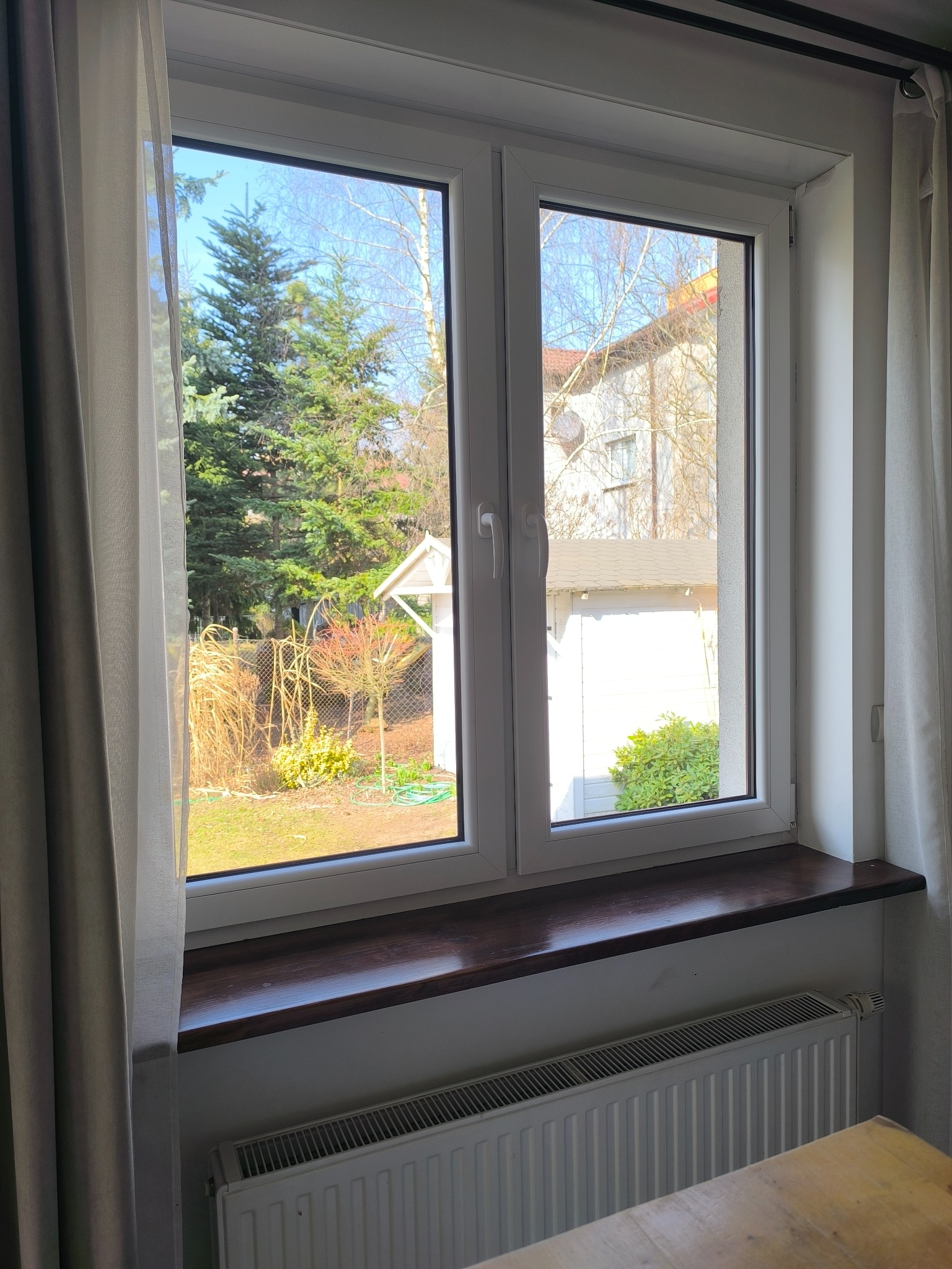 Białe okno z widokiem na ogród z iglakami i budynkiem w tle. Na parapecie widoczny grzejnik. Zasłona po lewej stronie okna.