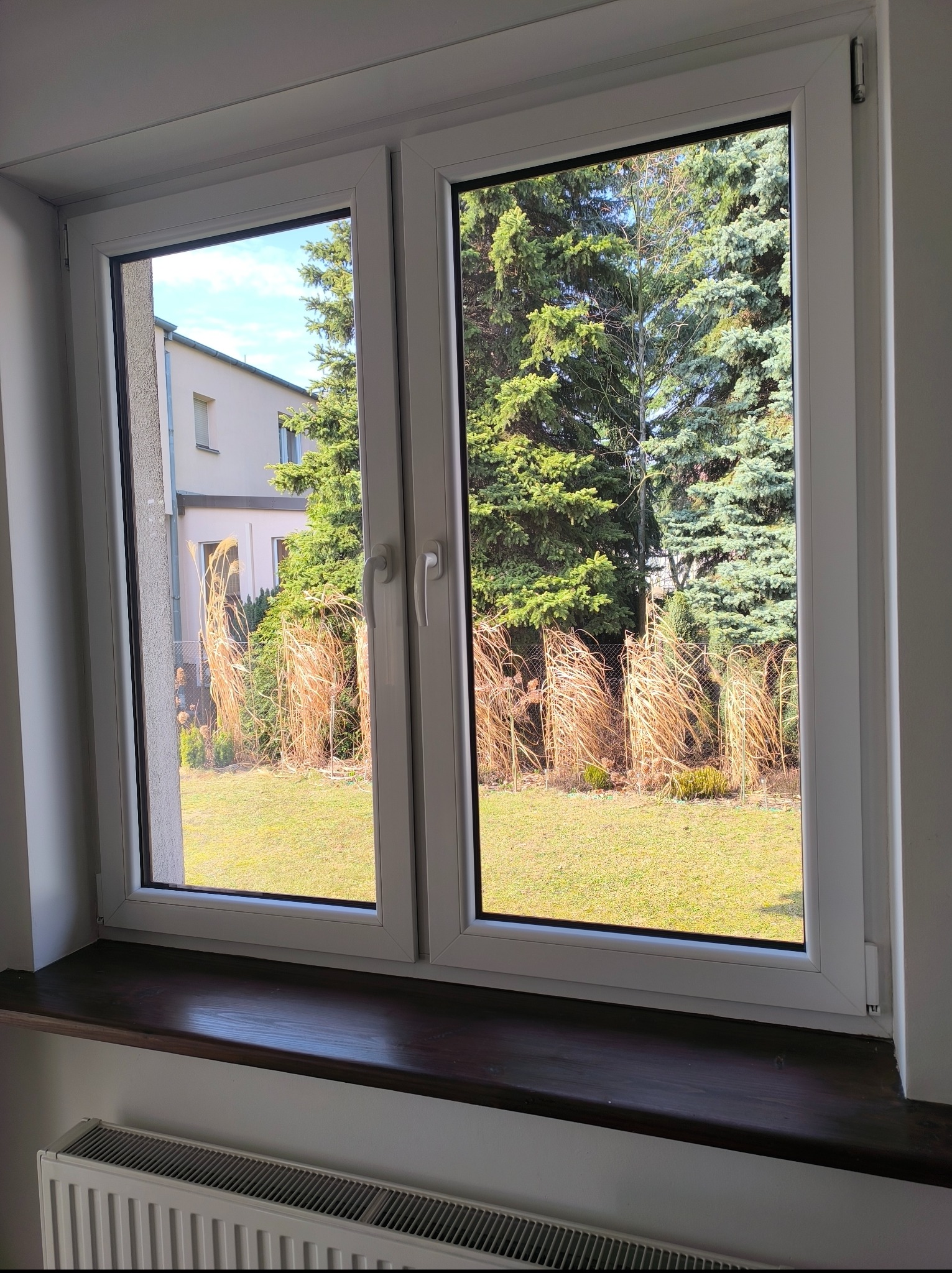 Białe, dwuskrzydłowe okno PCV z widokiem na zielony ogród z drzewami i ozdobnymi trawami. Na parapecie ciemne drewno, pod nim grzejnik.