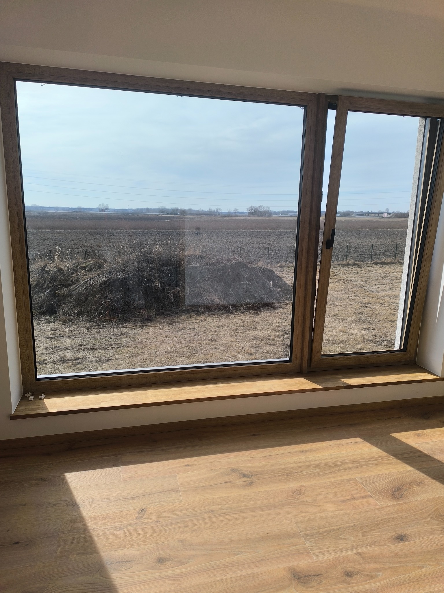 Drewniane okno z widokiem na pole i niebo. Częściowo otwarte skrzydło. Jasne panele podłogowe w pokoju. Słoneczny dzień.