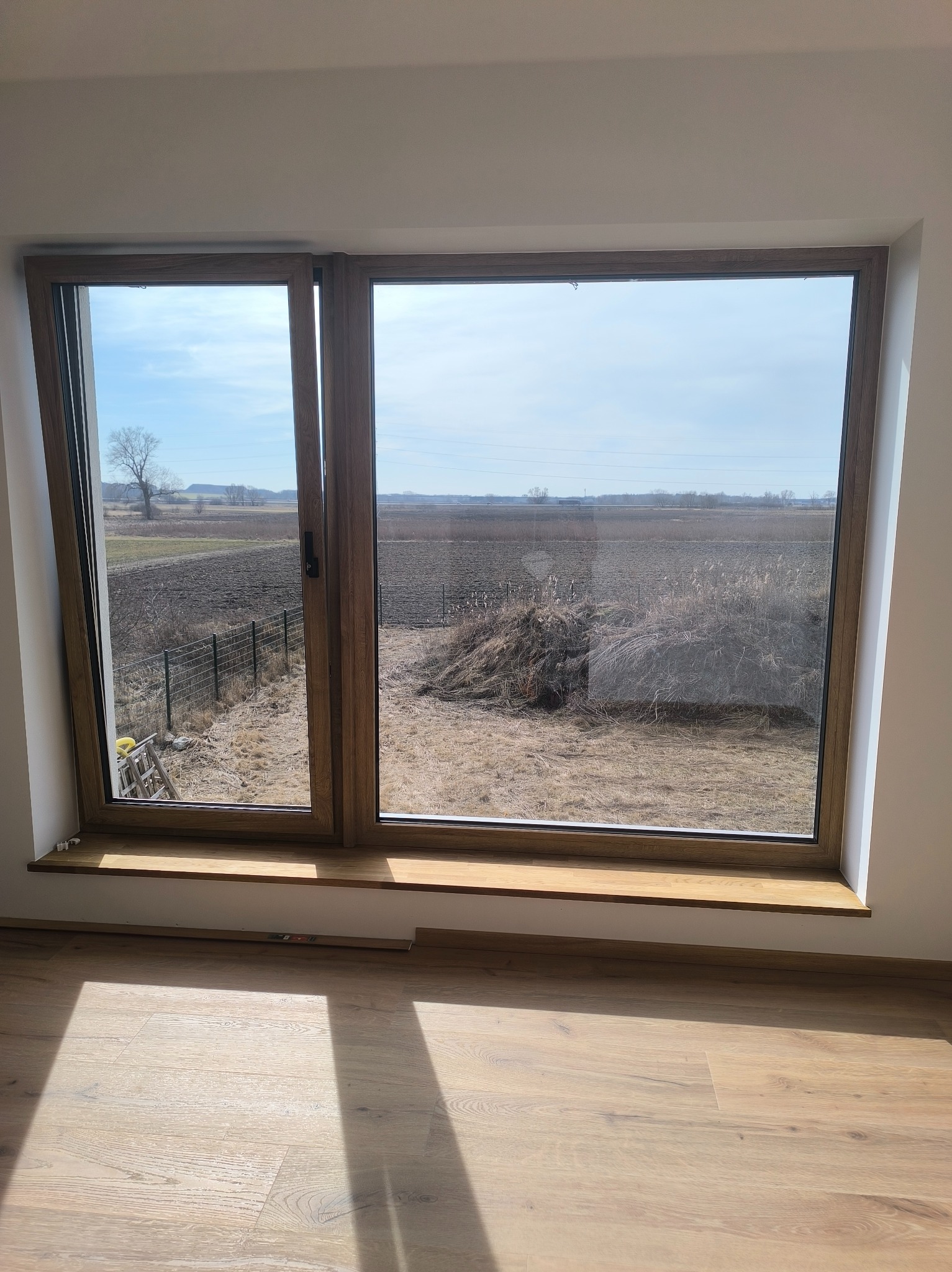 Duże okno z drewnianą ramą, widok na pole i niebo. Na parapecie leżą drobne elementy. Na podłodze widoczne promienie słoneczne.