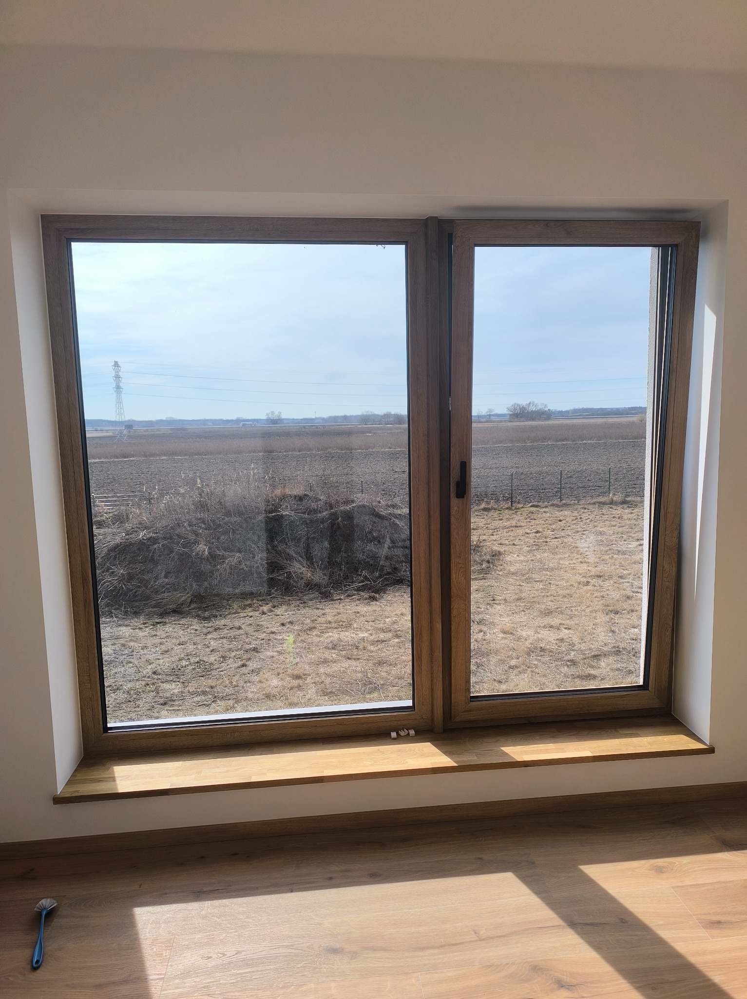 Drewniane okno z widokiem na pole w słoneczny dzień. Na parapecie leży mała szczotka. Podłoga z paneli odbija promienie słoneczne.