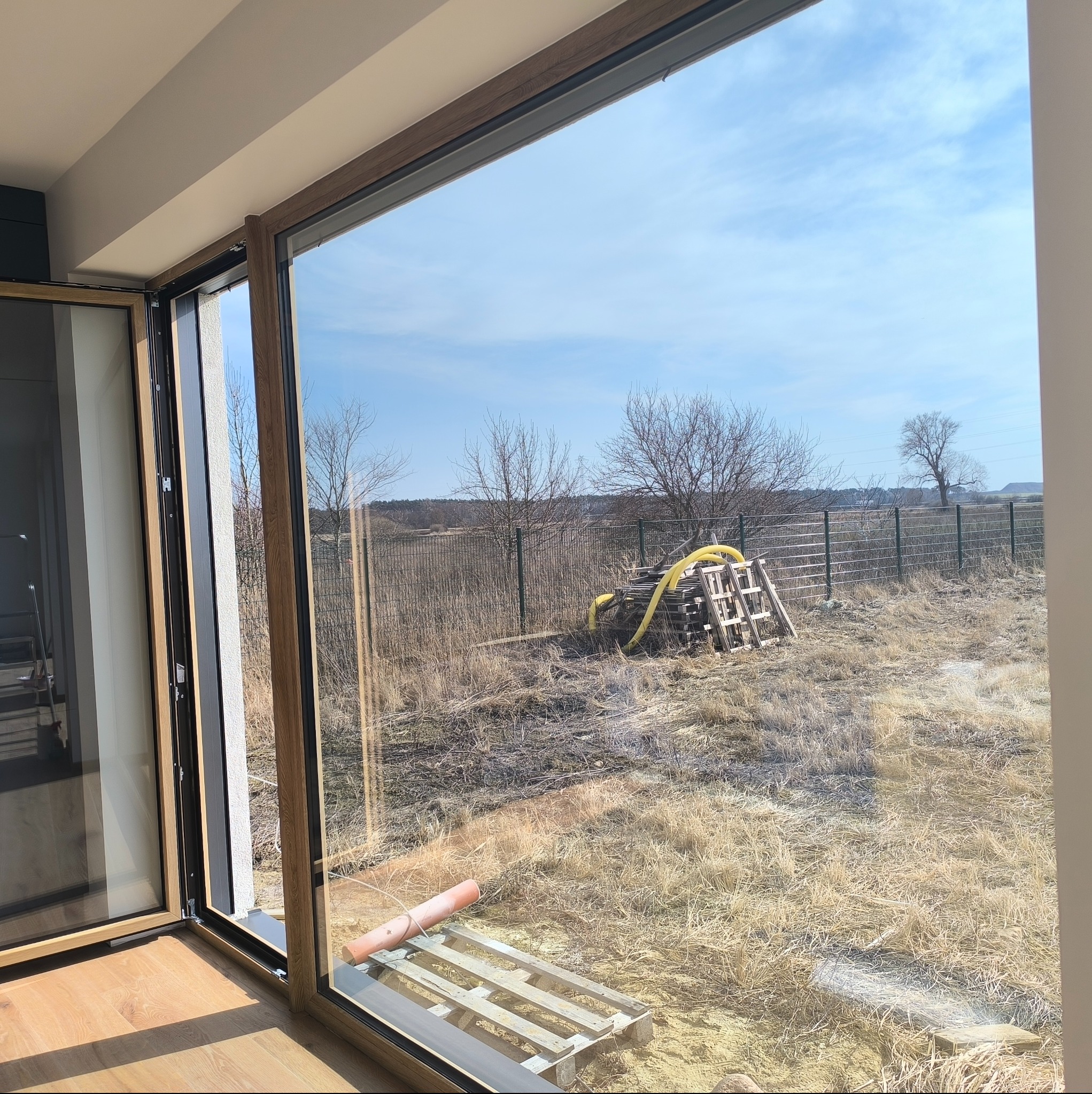 Otwarty drewniany portal okienny z widokiem na zaniedbane podwórko z paletami i rurami. Błękitne niebo w tle.