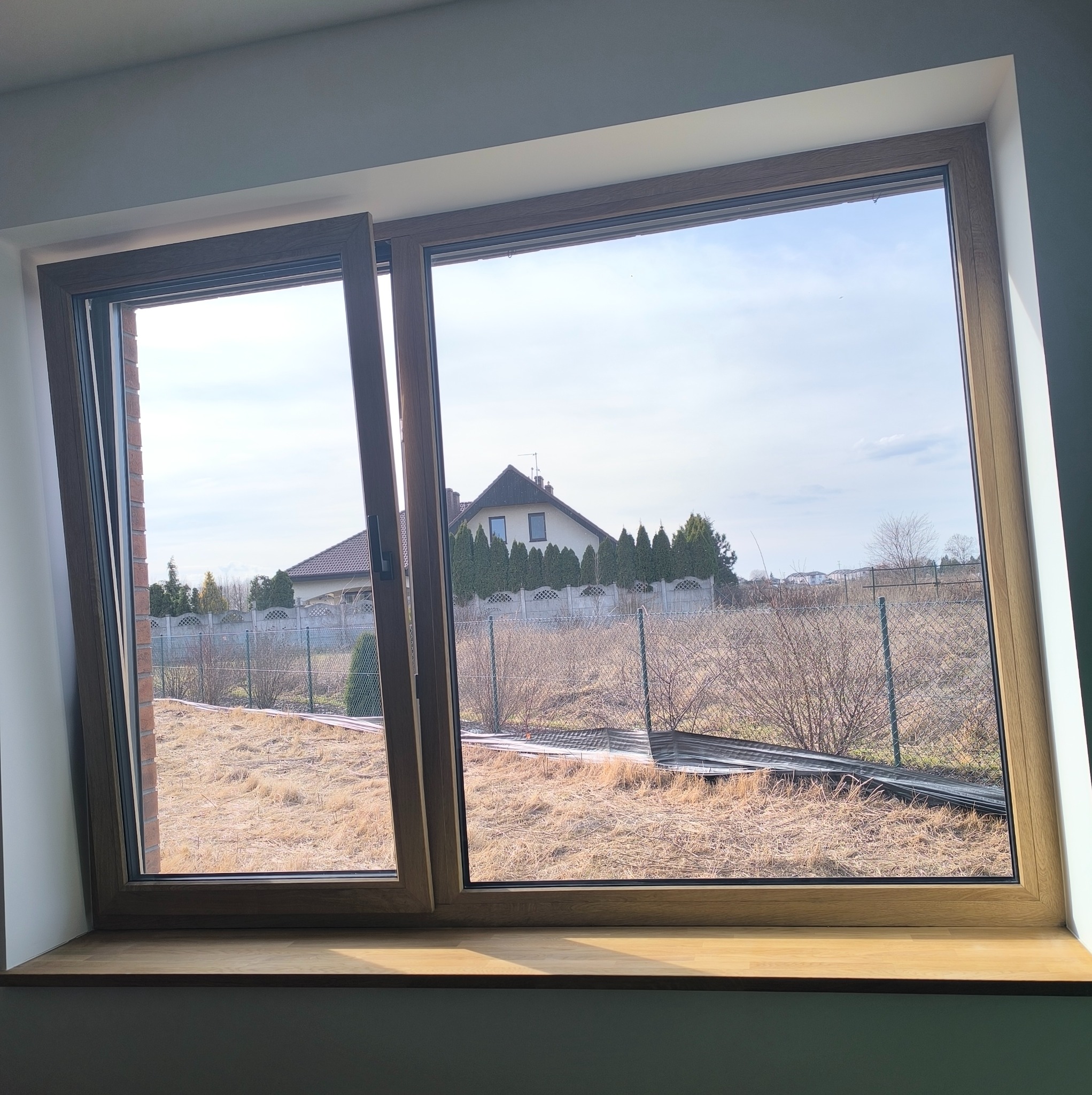 Drewniane okno z widokiem na ogród i dom. Jedna część okna jest uchylona, widać krajobraz za oknem w słoneczny dzień. Na parapecie widoczne refleksy światła.