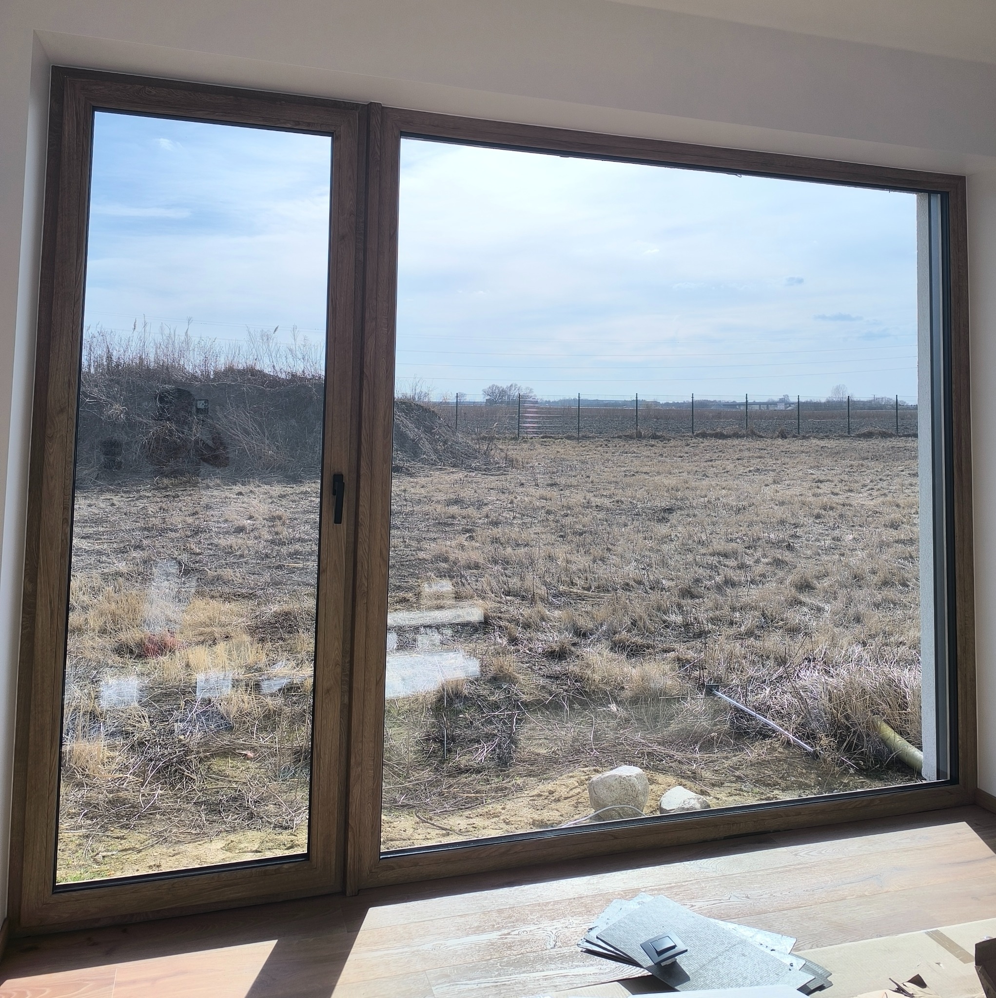 Duże okno z drewnianą ramą, widok na zewnątrz na pole z suchą trawą i ogrodzenie. Na podłodze leżą próbki płytek. Widok po umyciu okna.