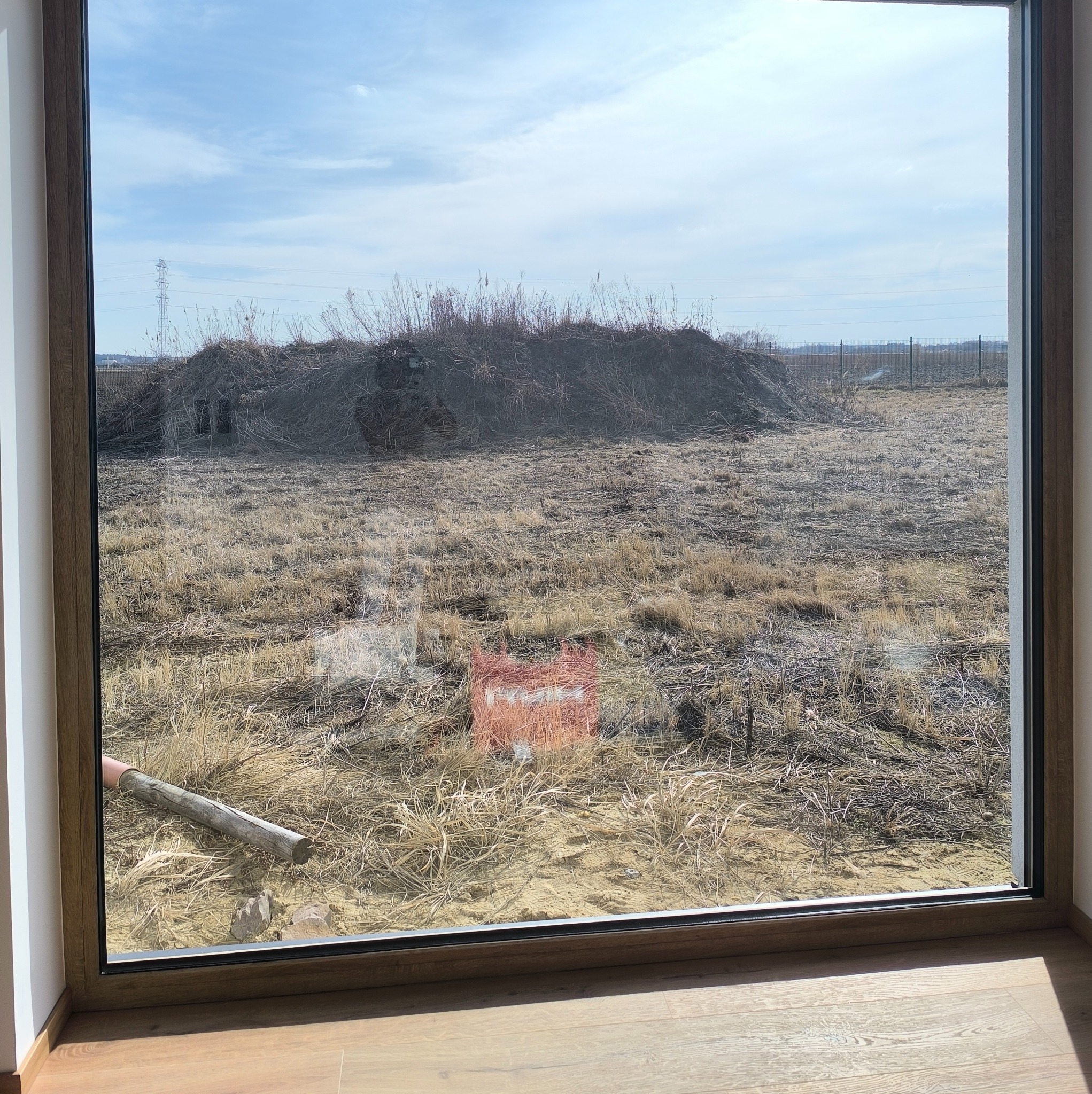 Widok przez okno na zaniedbane pole z uschniętą trawą i stertą gałęzi. W tle słupy wysokiego napięcia i ogrodzenie. Na pierwszym planie fragment drewnianej podłogi.