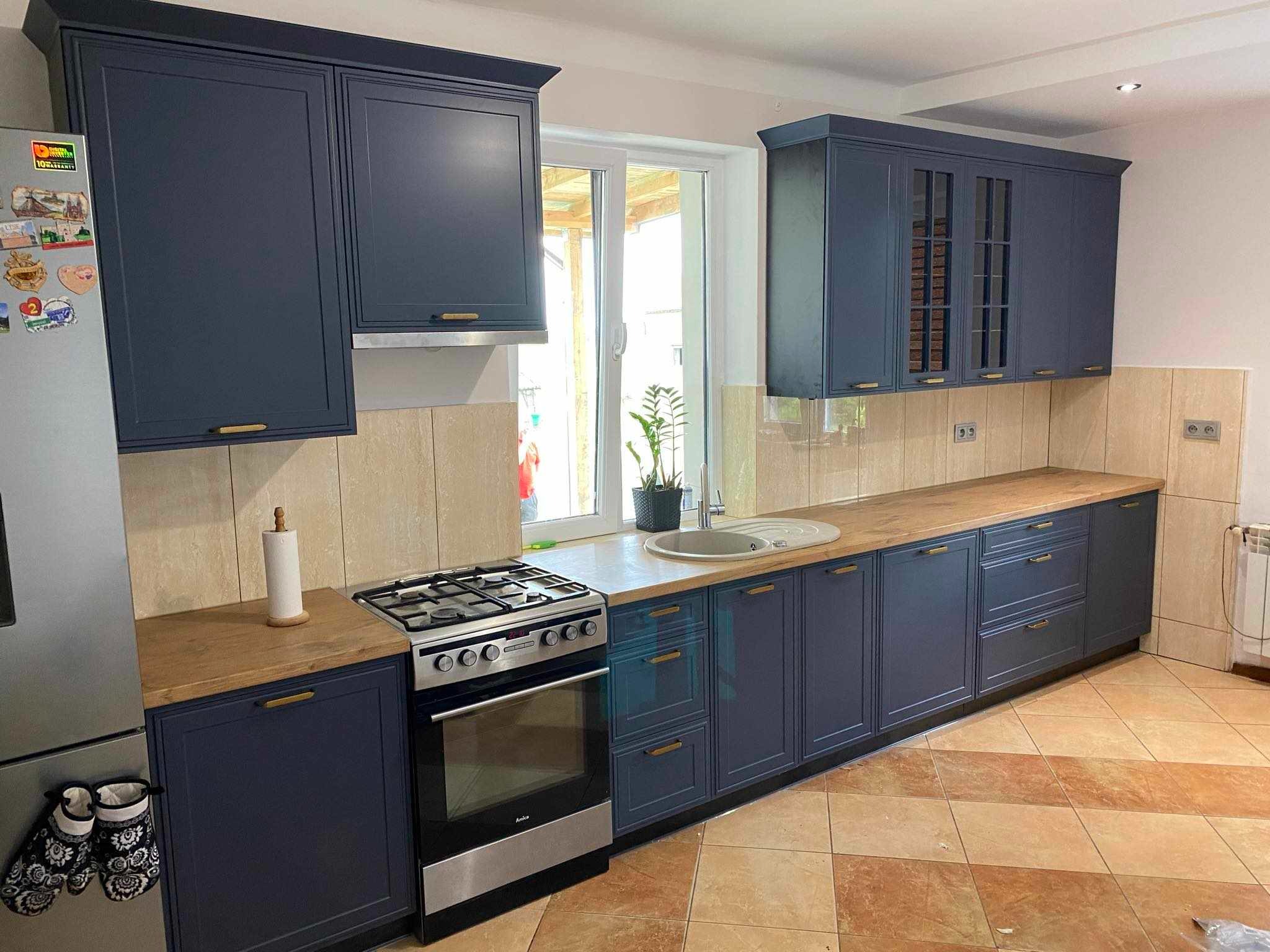 Granatowa zabudowa kuchenna z drewnianym blatem, widok na okno z rośliną. Górne szafki z przeszkleniami. Na lodówce magnesy. Płytki podłogowe w odcieniach brązu.