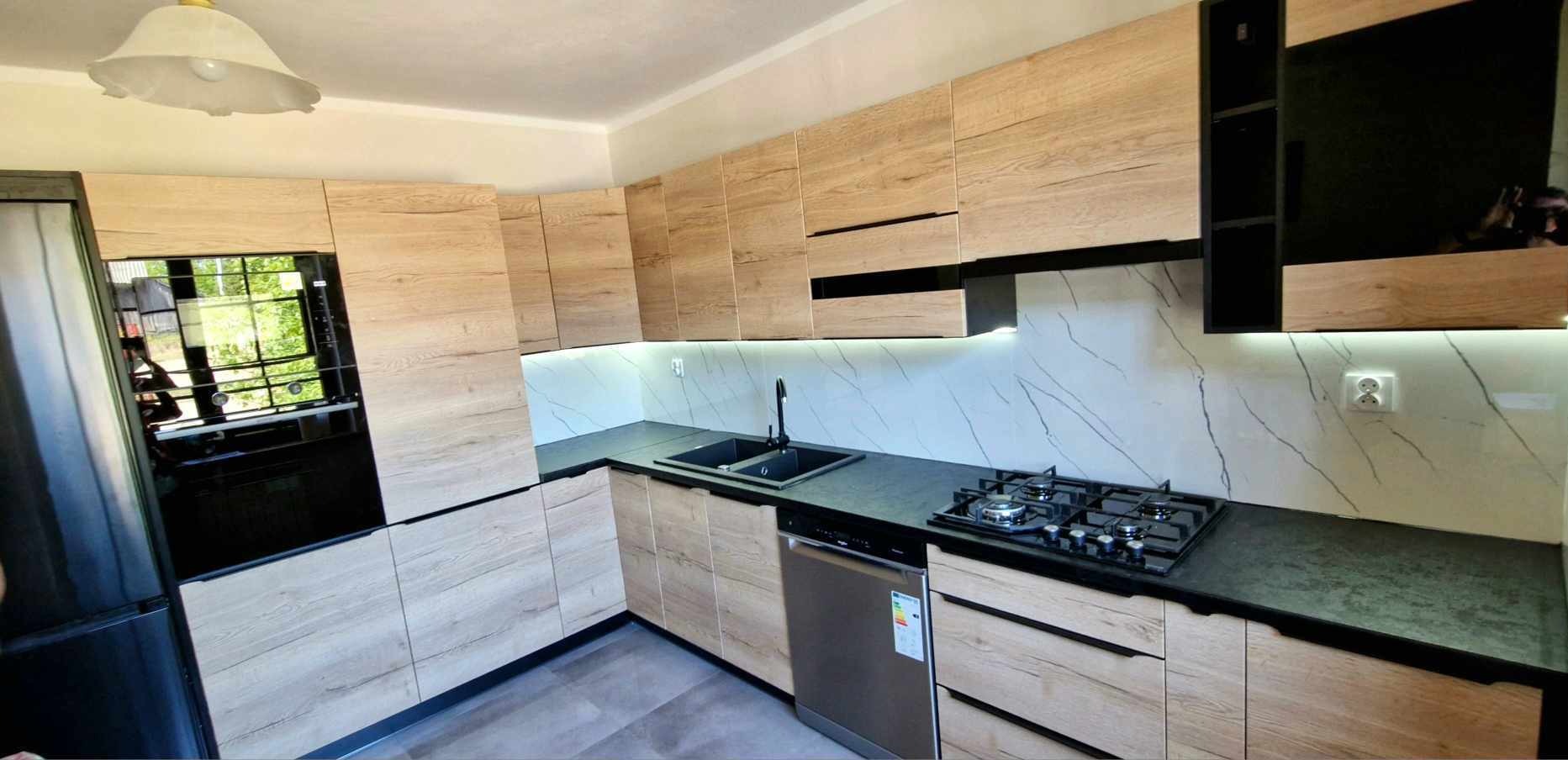 Jasna kuchnia na wymiar z drewnianymi frontami i czarnymi akcentami. Widoczny zlew, płyta gazowa i oświetlenie LED pod szafkami. Marmurowy wzór na ścianie.