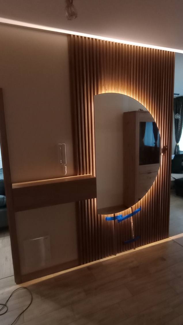 Nowoczesna zabudowa z drewnianych listew z okrągłym lustrem podświetlonym LED, widoczne elementy montażowe, ciepłe oświetlenie, minimalistyczny design.