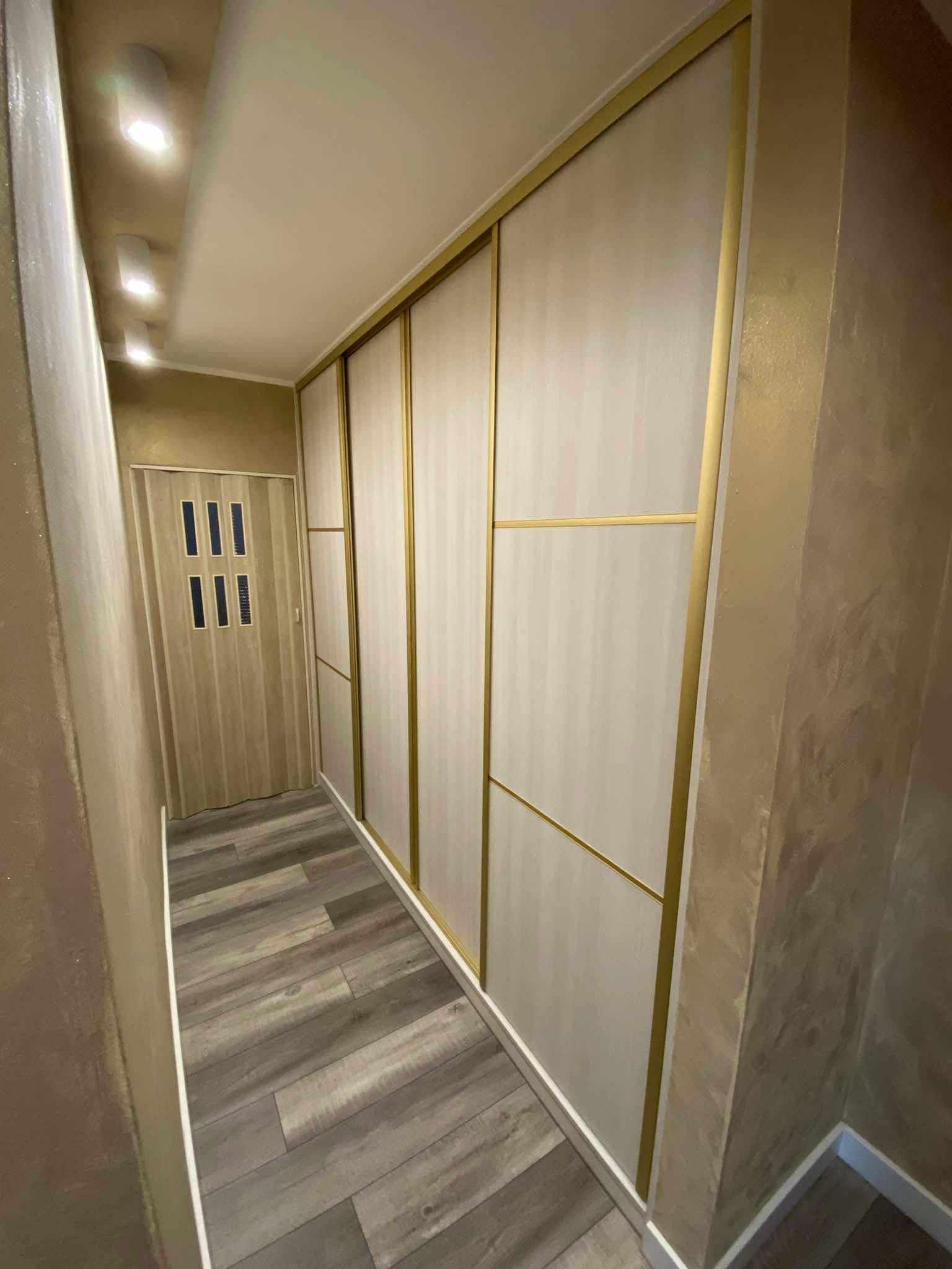 Biało-złota szafa przesuwna z drewnianymi panelami w wąskim korytarzu z panelami podłogowymi i dekoracyjną ścianą. Widoczna składana drewniana ścianka działowa.