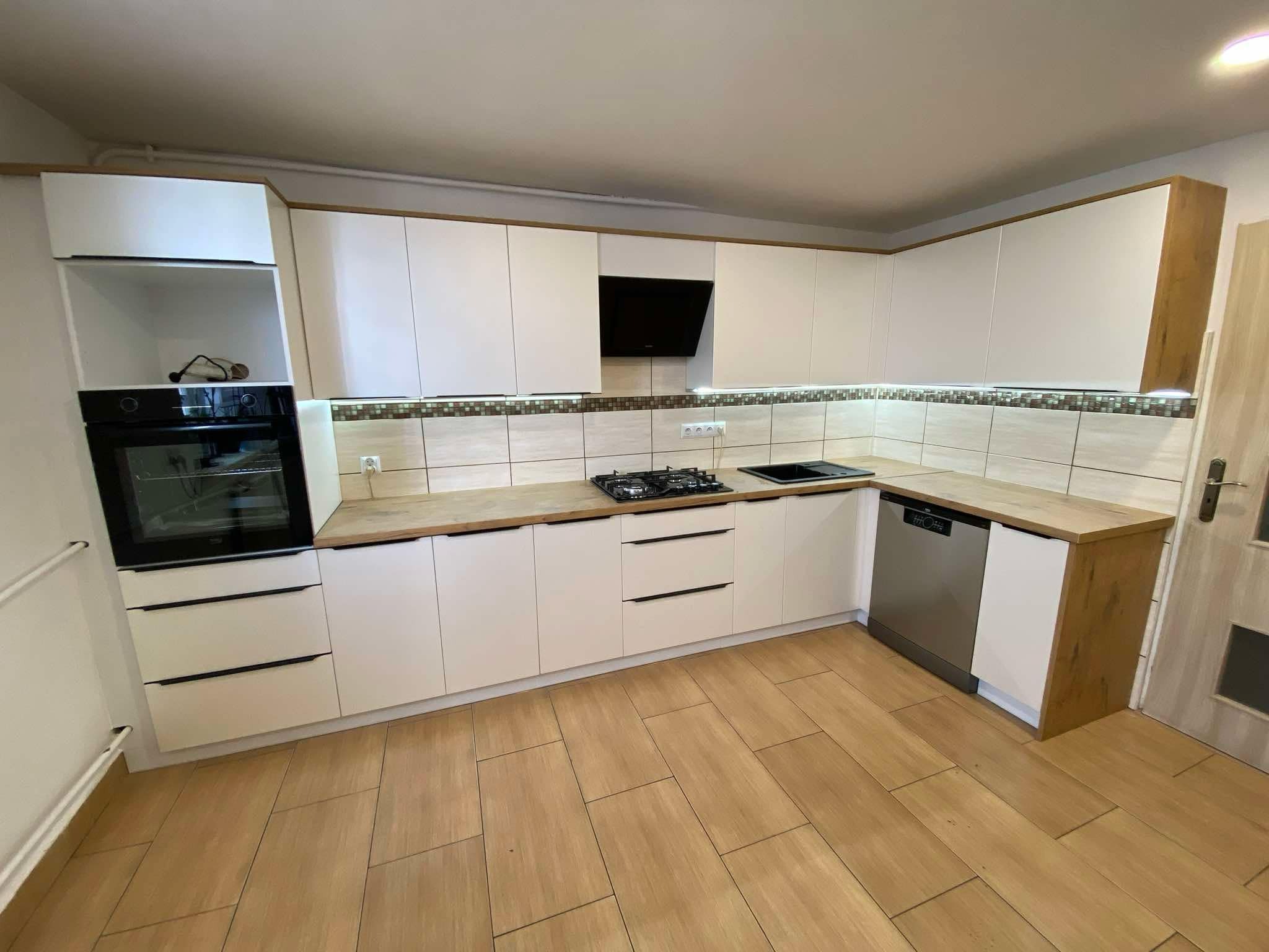 Jasna kuchnia z białymi szafkami i drewnianymi blatami. Uwagę przykuwa nowoczesny okap i płytki dekoracyjne nad blatem. Widoczna zabudowa piekarnika i zmywarki.